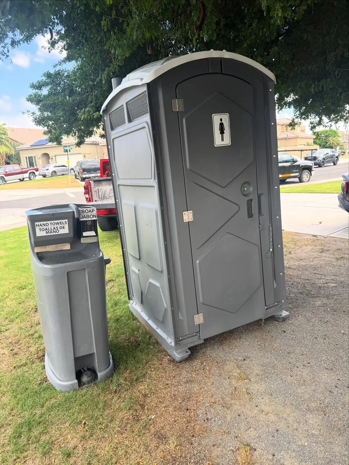 A gray portable toilet next to a hand wash station on a green lawn
